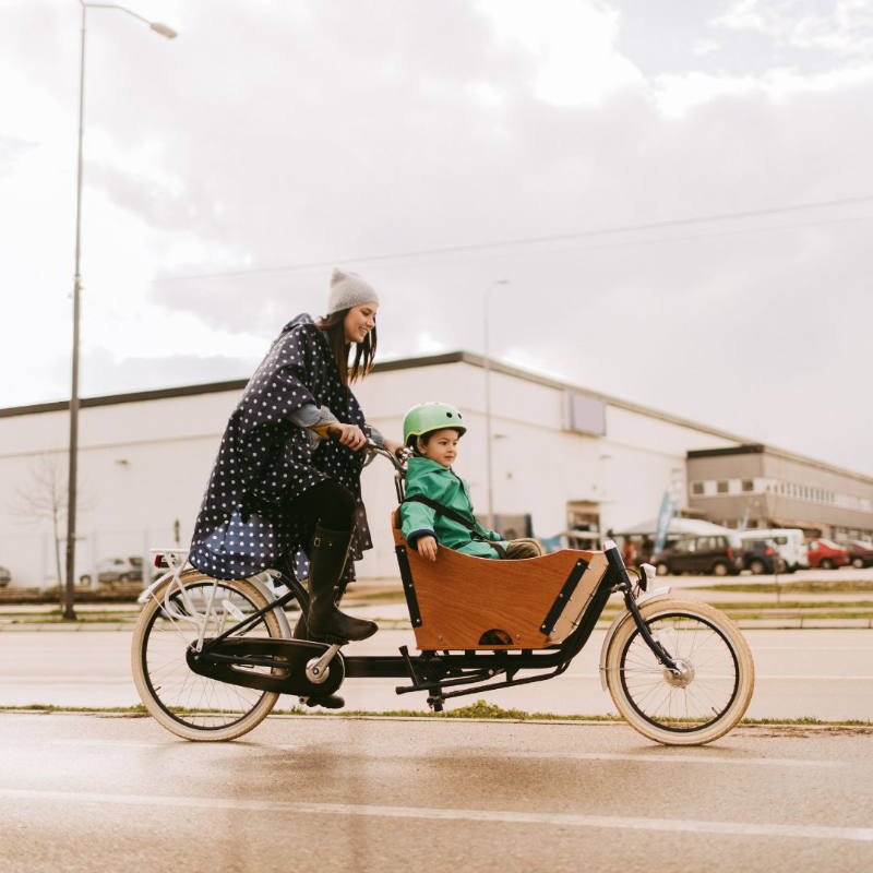 weiblich gelesene Person fährt mit Kind mit Lastenrad auf der Straße